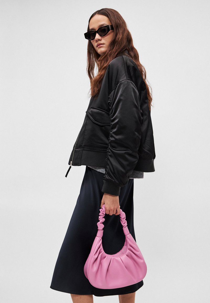 Sort satin bomberjakke med rynkede ærmer, kombineret med en sort satin nederdel. Holder en blød lyserød plisseret håndtaske med en snoet strop.