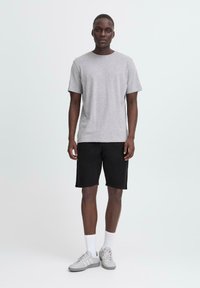 Camiseta gris de manga corta hecha de algodón, combinada con pantalones cortos negros. El modelo lleva zapatillas grises y calcetines blancos, de pie contra un fondo simple.