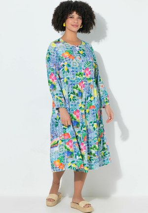 Frau trägt ein farbenfrohes, gemustertes Midikleid mit langen Ärmeln und gelbe Creolen, steht vor einem weißen Hintergrund.