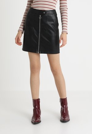 Femme portant une mini-jupe en simili cuir noir avec une fermeture éclair devant, un haut à manches longues à rayures, et des bottines brillantes rouge foncé, debout sur un sol blanc.