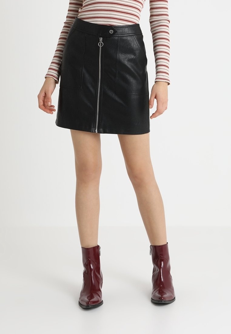 Femme portant une mini-jupe en simili cuir noir avec une fermeture éclair devant, un haut à manches longues à rayures, et des bottines brillantes rouge foncé, debout sur un sol blanc.