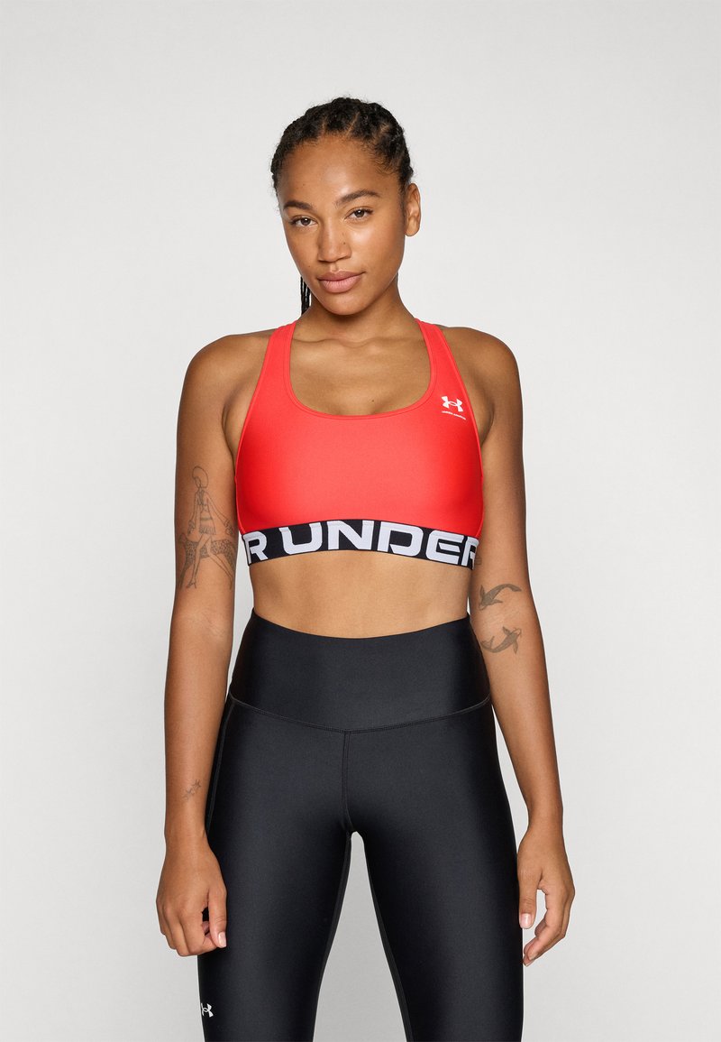 Sostén deportivo rojo con una banda ancha negra que presenta la palabra "UNDER" en blanco. La tela es suave y ajustada, combinada con leggings negros de cintura alta.