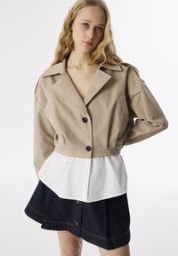 Beige verkürzte Jacke mit einem weißen Saum, featuring einem Reverskragen und zwei Knöpfen. Kombiniert mit einem dunkelblauen Denimrock mit goldenen Nähten.