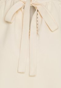 Blusa color crema con chiusura a fiocco al collo, caratterizzata da delicati accenti a scallop e tessuto morbido e liscio.