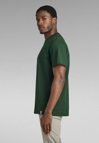T-shirt in cotone verde scuro a maniche corte con scollatura rotonda. Presenta un'etichetta sull'orlo sinistro e una vestibilità standard. Texture liscia.