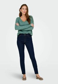 Donna in piedi con le braccia incrociate, che indossa un maglione verde, jeans blu scuri e scarpe basse marroni, su uno sfondo chiaro e neutro.