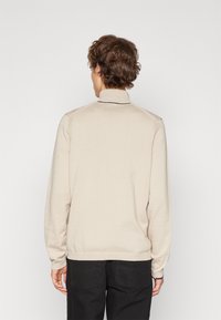 Pull beige avec un col montant, texture tricotée et accents au niveau des coutures d'épaule. Manches et ourlet côtelés. Vue de dos avec un minimum de détails.