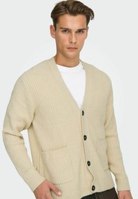 Cremfarbener gerippter Cardigan aus Strickstoff, mit V-Ausschnitt, Frontknöpfen und zwei offenen Taschen. Strukturierte, weiche Optik.
