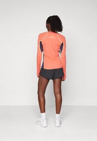 Langærmet atletisk top i koralfarve med blå detaljer, har en tætsiddende pasform og kontrasterende syninger, parret med sorte atletiske shorts.