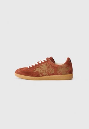 Zapatilla de ante color marrón óxido con cordones blancos, suela de goma, motivo floral en el lateral y firma "Paul Smith" en letra dorada.