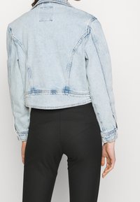 Žena v krátké světle modré denimové bundě s tmavým prošíváním, v kombinaci s těsnými černými kalhotami, zobrazená zezadu.