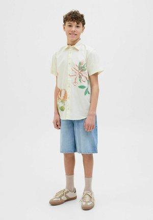 Chemise jaune à manches courtes avec imprimé floral, associée à un short en denim et des baskets marron clair. Des chaussettes beiges complètent le look.