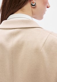 Jasne ružové sako s textúrovaným diamantovým vzorom; obsahuje klasický golier a jediný šev. Viditeľný je veľký strieborný kruhový náušník.