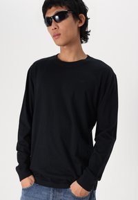Tricou negru cu mâneci lungi, fabricat din bumbac, design cu guler rotund, având o croială lejeră. Modelul poartă ochelari de soare negri și blugi albaștri.