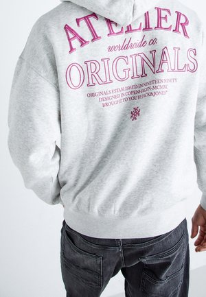Osoba nosząca jasnoszarą bluzę z kapturem z różowym napisem "Atelier Originals" i ciemne dżinsy, widoczna od tyłu na jednolitym tle.