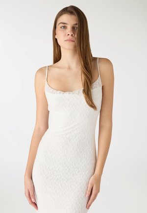 Giovane donna con lunghi capelli castani che indossa un vestito bianco senza maniche in pizzo, in piedi davanti a uno sfondo chiaro e uniforme.