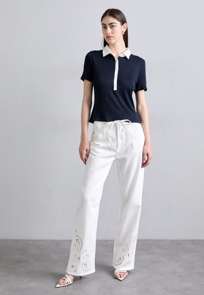 Navy short-sleeve polo with a white collar, paired with white wide-leg pants featuring floral eyelet cutouts at the hem; worn with white heels.