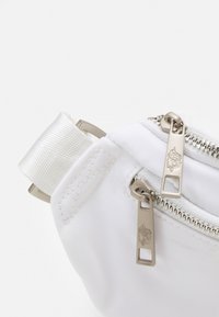 Zbliżenie rogu białej torby materiałowej z dwoma srebrnymi zamkami błyskawicznymi, na których znajdują się kwiatowe grawerunki na uchwytach oraz przyszyta pętelka.