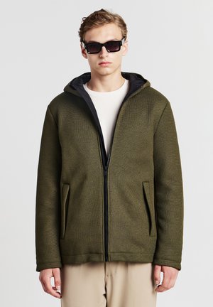 Giacca verde oliva con tessuto strutturato, zip nera e cappuccio. Presenta tasche laterali e una vestibilità comoda, abbinata a una camicia bianca sotto.
