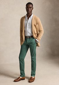 Homme portant un cardigan beige, une chemise blanche boutonnée, un pantalon vert, une ceinture marron, des mocassins marron et des lunettes, debout avec une main dans la poche.