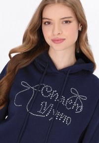 Sudadera con capucha azul marino con cordón, con un diseño hecho de brillantes plateados. El material parece suave, con un ajuste relajado.