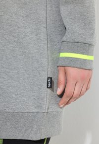 Sudadera gris con cuello redondo, puños acanalados y una franja de acento amarilla brillante en la muñeca. Presenta una etiqueta de logo "sprts" en el lateral.