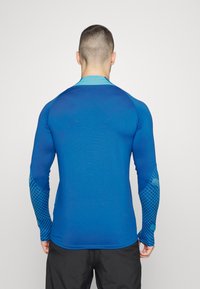 Sininen urheilu pitkähihaista paitaa, jossa on gradienttikuviot, teksturoidut hihnat ja pyöreä kaula-aukko. Kangas näyttää sileältä ja istuvalta.