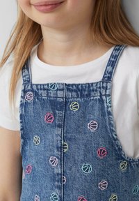 Denim overall s pisanim večbarvnim vzorcem školjk na sprednji strani, z nastavljivimi naramnicami in belim majico z dolgimi rokavi spodaj.