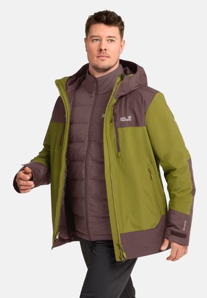 Mann trägt eine grün-braune Jack Wolfskin Outdoorjacke über einer braunen Daunenjacke und geht mit neutralem Gesichtsausdruck.