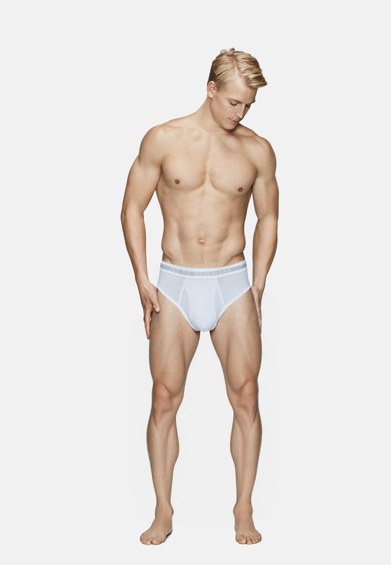 Hvide bomuldsbriefs med et mærket elastikbånd, der har en tætsiddende pasform og en glat tekstur. Modellen har en slank, muskuløs kropsbygning.