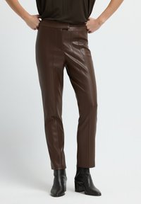 Bruine faux leren broek met een getailleerde silhouette, voorzien van verticale naden en een gladde textuur. Gecombineerd met zwarte hoge enkellaarsjes.