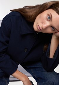 Cappotto in misto lana blu navy con colletto appuntito, bottoni frontali e maniche ampie. Presenta una texture liscia e una vestibilità rilassata.