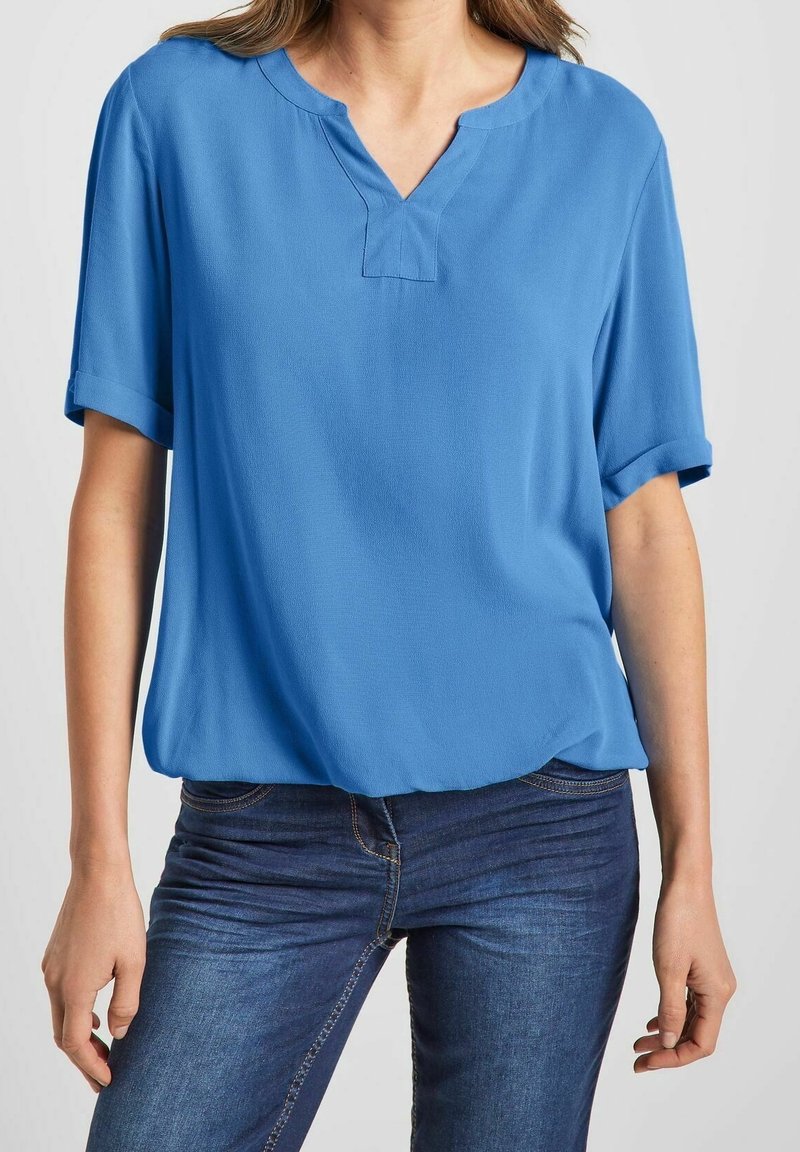 Femme portant un chemisier bleu ample à manches courtes avec un décolleté en forme d'encoche, associé à un jean bleu foncé.