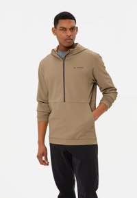 Hellbraunes Kapuzensweatshirt mit einem halbseitigen Reißverschluss, strukturiertem Stoff und einem Logo auf der Brust. Kombiniert mit schwarzen Sporthosen.