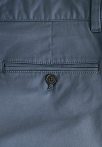 Tasca posteriore di pantaloni blu con chiusura a un solo bottone e passante per cintura sopra.