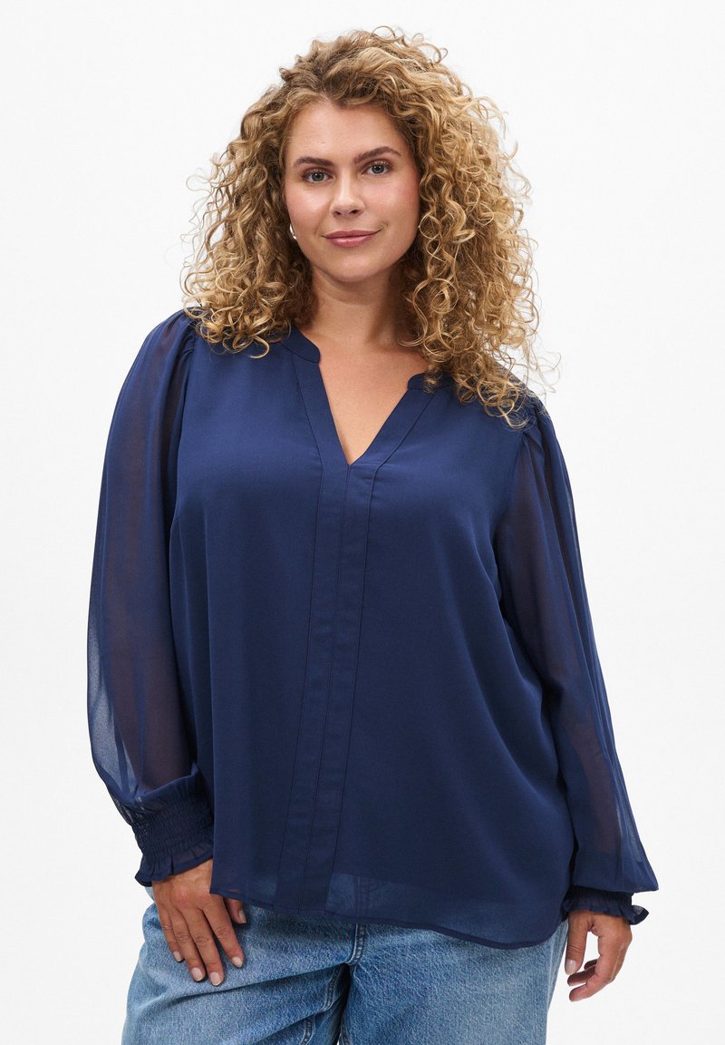 Blouse bleu marine avec un col en V, des manches longues en voile et des poignets froncés. Tissu léger avec une texture subtile et une coupe décontractée.