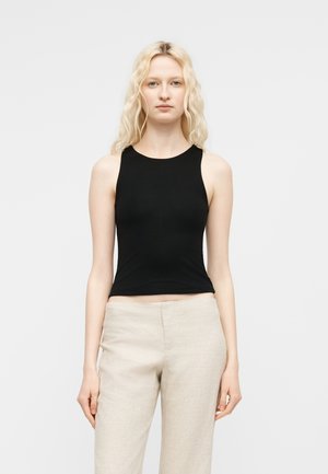 Γυναίκα με ανοιχτόξανθα μακριά μαλλιά φοράει αμάνικη μαύρη μπλούζα και ανοιχτό μπεζ παντελόνι, στέκεται μπροστά σε ένα απλό λευκό φόντο.