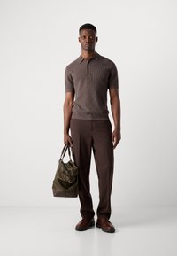 Camisa de polo marrón de manga corta con textura y tres botones, combinada con pantalones marrones. Sosteniendo un bolso de cuero verde oliva con un cierre ajustable.