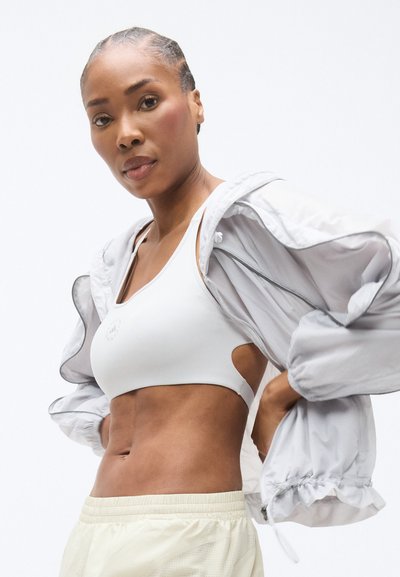 Λευκό αθλητικό σουτιέν με κοψίματα στα πλαϊνά, γκρι ελαφρύ μπουφάν με ελαστικές μανσέτες και ανοιχτό μπεζ σορτς με υφασματικό μοτίβο.