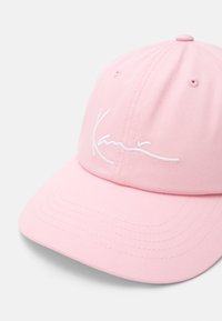 Pink baseball cap made of cotton, featuring a curved brim and white embroidered logo on the front. Subtle ribbed texture throughout.