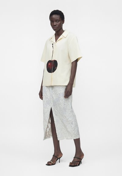 Γυναίκα που φοράει μια κρεμ κοντομάνικη μπλούζα με μεγάλο γραφικό κεράσι και μια λευκή δαντελένια μίντι φούστα με ανοίγματα εμπρόσθια, στέκεται με μαύρα σανδάλια με τακούνι.