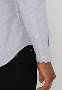 Vaaleanharmaa pitkähihainen paita, jossa on valkoiset napit, käytetään mustien farkkujen kanssa, ja näkyvissä ovat sivusauman ja takataskun yksityiskohdat.