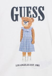 Γραφικό με μια αρκούδα που φορά ένα μπλε φόρεμα με λουλούδια και βολάν, λευκά αθλητικά παπούτσια, και κείμενο που γράφει "GUESS" και "LOS ANGELES EST. 1981."