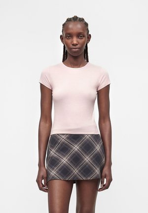 Kvinde med fletninger har en lyserød tætsiddende T-shirt på og et kort mørkt ternet skørt, stående foran en ensfarvet hvid baggrund.