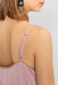 Donna che indossa un top a spaghetto color malva con spalline regolabili, mostrando la spalla, parte del viso e un orecchino floreale nero.