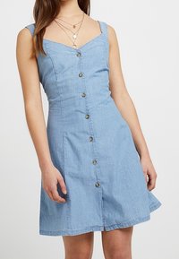 Ljust blå denimklänning med en figurnära överdel, nylonband och en utsvängd kjol. Den har knäppning fram med knappar i guldton.