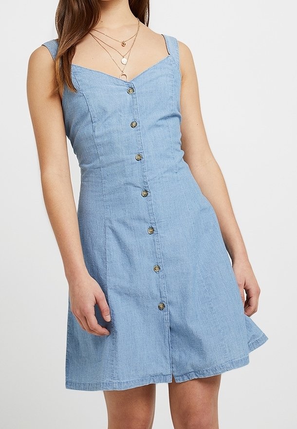 Ljust blå denimklänning med en figurnära överdel, nylonband och en utsvängd kjol. Den har knäppning fram med knappar i guldton.