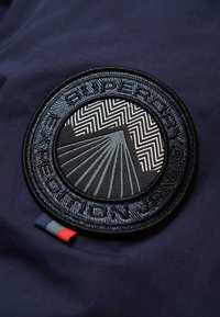 Parche circular de tela negra con diseño en relieve de zigzag y rayos de sol, texto "SUPERDRY EXPEDITION JAPAN", sobre un fondo azul marino oscuro.