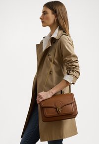 Brown leather handbag with a structured design, featuring a gold accent clasp and a chain strap, held by a person wearing a beige trench coat.