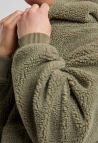 Personne ajustant le col d'une veste polaire verte texturée avec des poignets côtelés visibles aux manches.